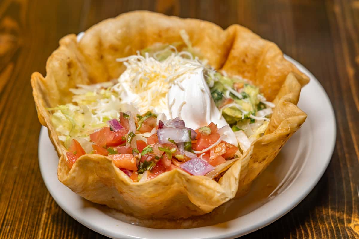 Crispy taco salad bowl filled with lettuce, guacamole, pico de gallo, shredded cheese, and sour cream on a white plate.
