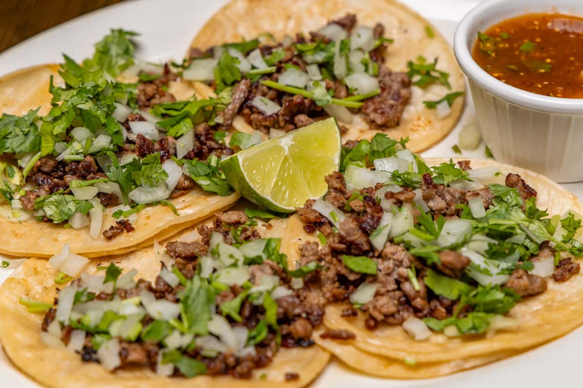 Street-style carne asada tacos topped with chopped onions and cilantro, served with lime wedges and a side of salsa.