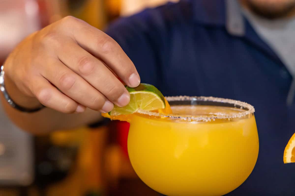 Hand placing a lime and orange garnish on a salt-rimmed mango margarita in a goblet glass.