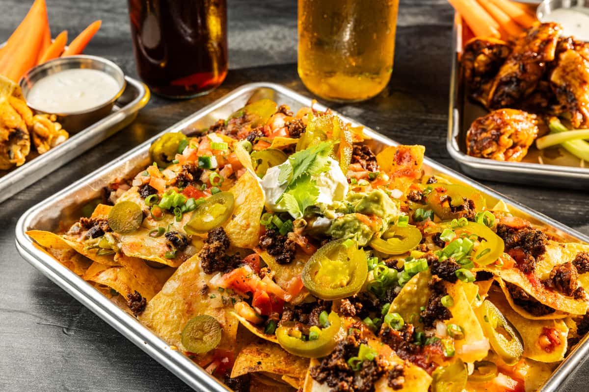 A tray of nachos topped with seasoned ground beef, jalapeños, green onions, sour cream, guacamole, and diced tomatoes, with drinks in the background.