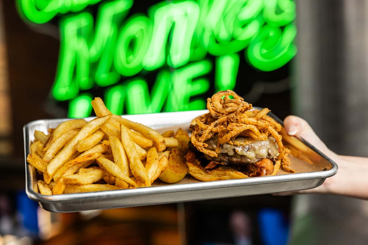 A person holds a tray with a cheeseburger topped with crispy onions and served with a side of French fries, set against a neon "Gas Monkey Live!" sign in the background.