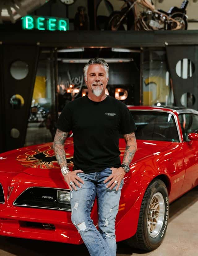 A man with tattoos and a goatee poses in front of a red Pontiac Firebird Trans Am, standing in a garage with a neon "BEER" sign.