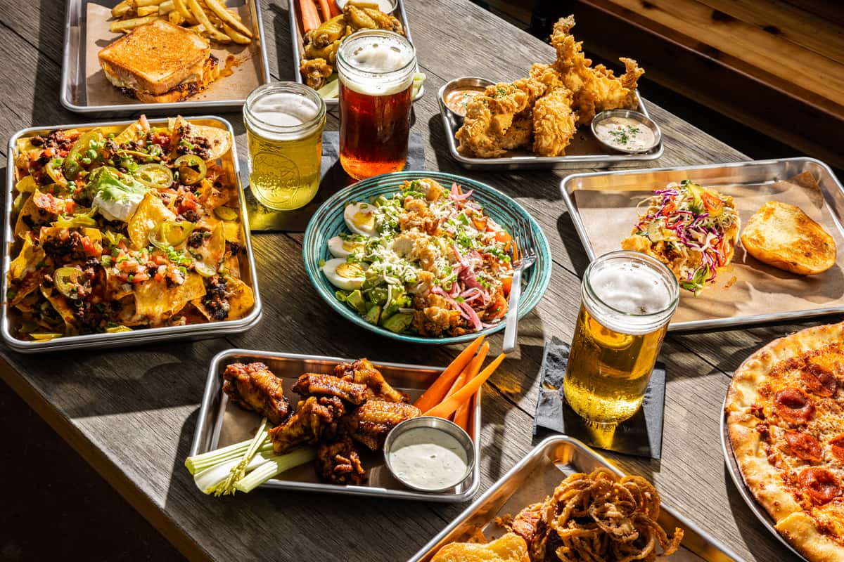 A spread of food and drinks, including nachos, fried chicken, a salad, wings with celery, a sandwich, pizza, and beers on the table.