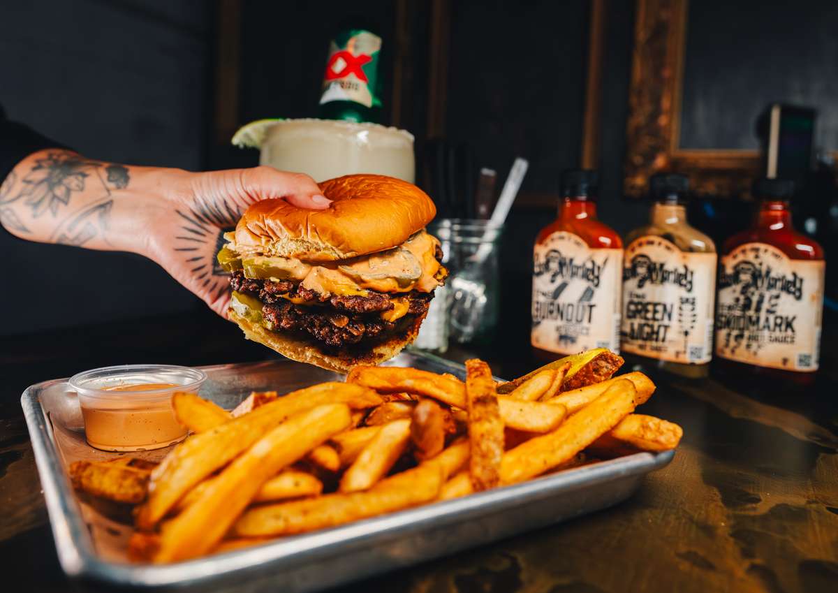 A hand holds a large burger with two patties, pickles, and sauce, accompanied by a side of fries and bottles of hot sauce.