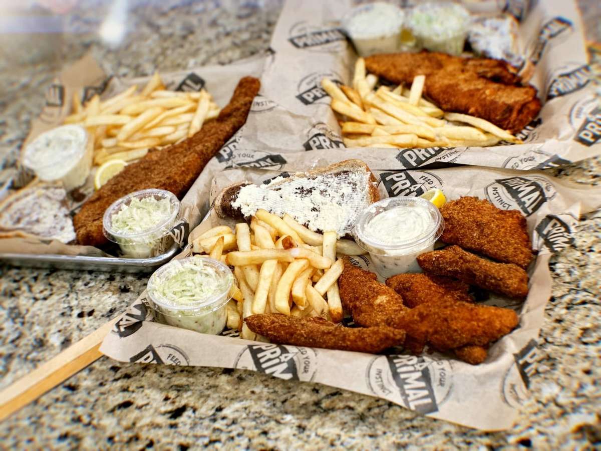 Classic Wisconsin Fish Fry with french fries, slaw, and a slice of lemon, on a platter with a paper layer with text: Primal Eats, Feed Your Instinct