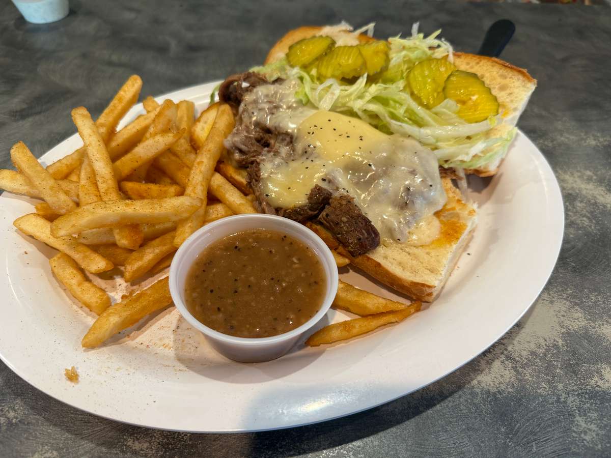 Open-faced roast beef sandwich topped with melted cheese, pickles, and shredded lettuce, served with seasoned French fries and a side of brown gravy.