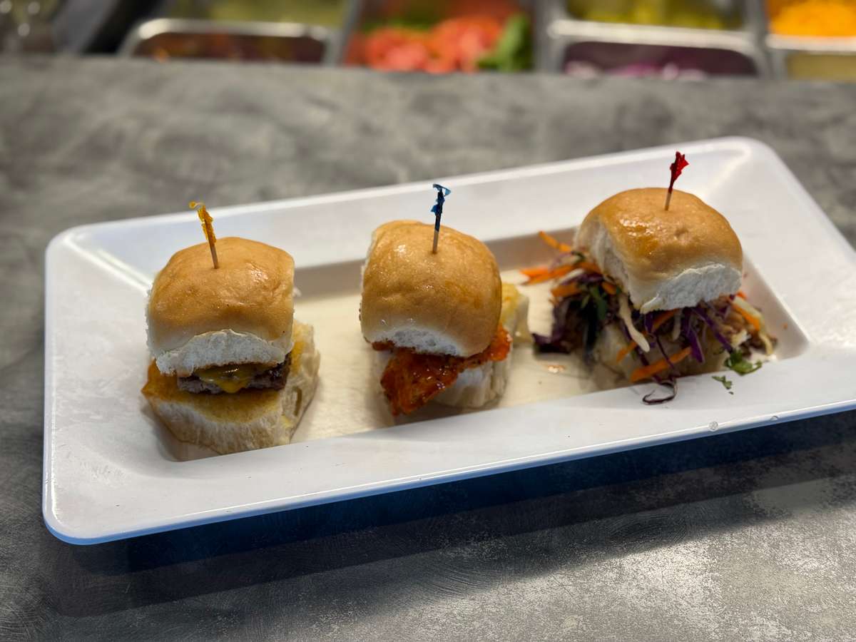 Trio of gourmet sliders on a white rectangular plate, including a cheeseburger, spicy chicken, and coleslaw-topped pulled pork slider.