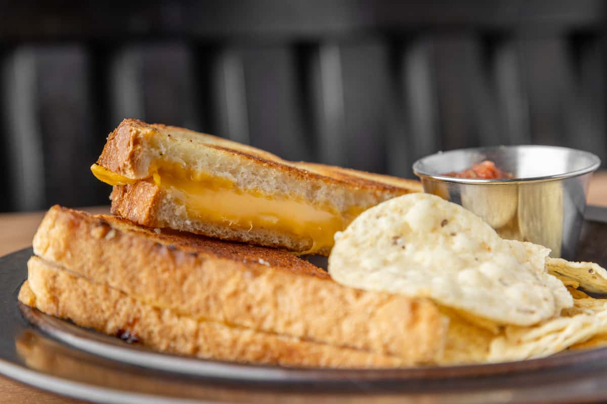 Grilled cheese sandwich with melted cheddar on toasted bread, served with kettle chips and a side of dipping sauce in a classic diner setting