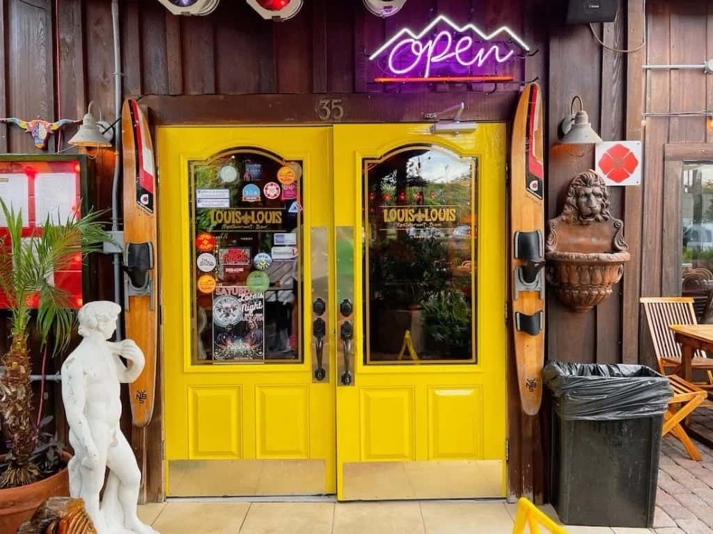 The entrance of "Louis Louis" restaurant with vibrant yellow double doors, a neon "Open" sign, and decorative elements like ski poles and a statue.