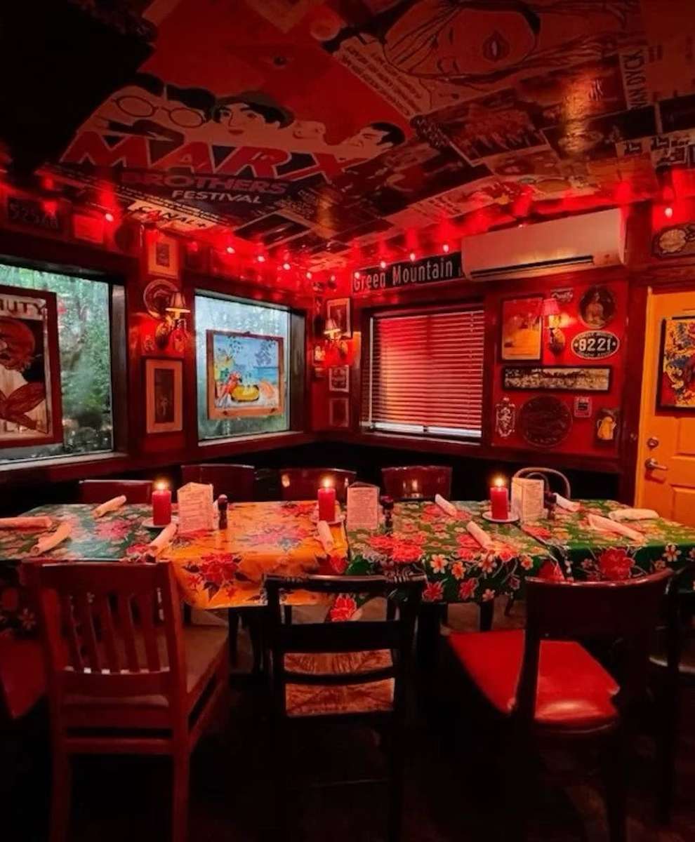 The cozy interior of a restaurant featuring red lighting, vintage decor, and a large floral tablecloth with candles placed at each setting.