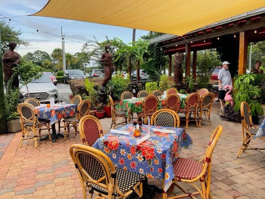 A vibrant outdoor seating area with colorful floral tablecloths, wicker chairs, potted plants, statues, and a relaxed atmosphere under a sunshade.