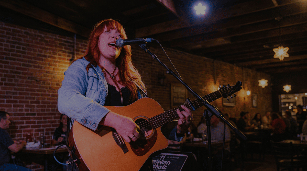 A woman with red hair sings into a microphone while playing an acoustic guitar on a dimly lit stage.