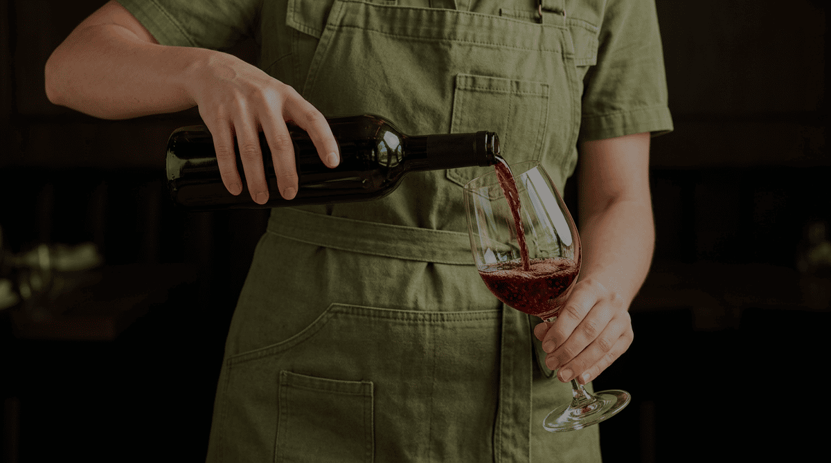 A person in green overalls pouring red wine from a dark bottle into a clear wine glass, held in their other hand.