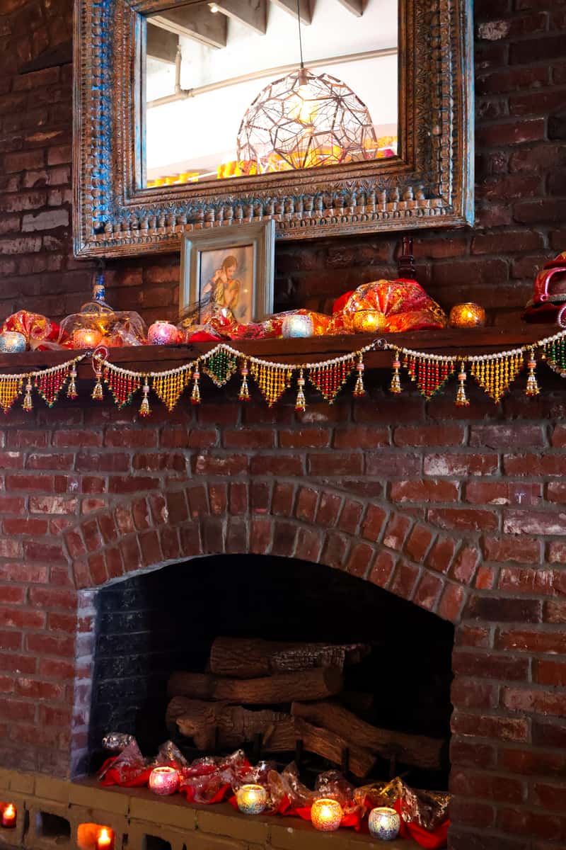 A decorated mantel with string lights, colorful fabric, and candles. A framed picture of a person is placed above, reflected in a large mirror.