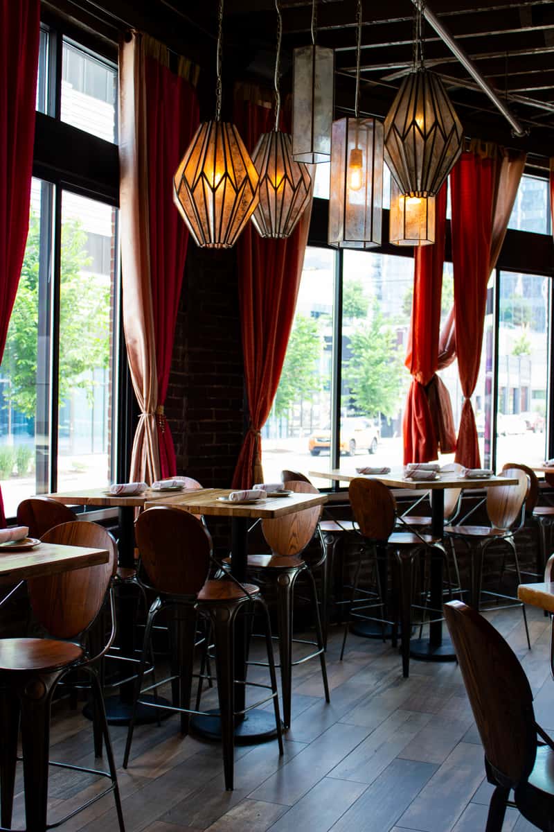 A cozy restaurant setting with wooden tables, chairs, and stylish pendant lights hanging above, while large windows allow natural light to fill the space.