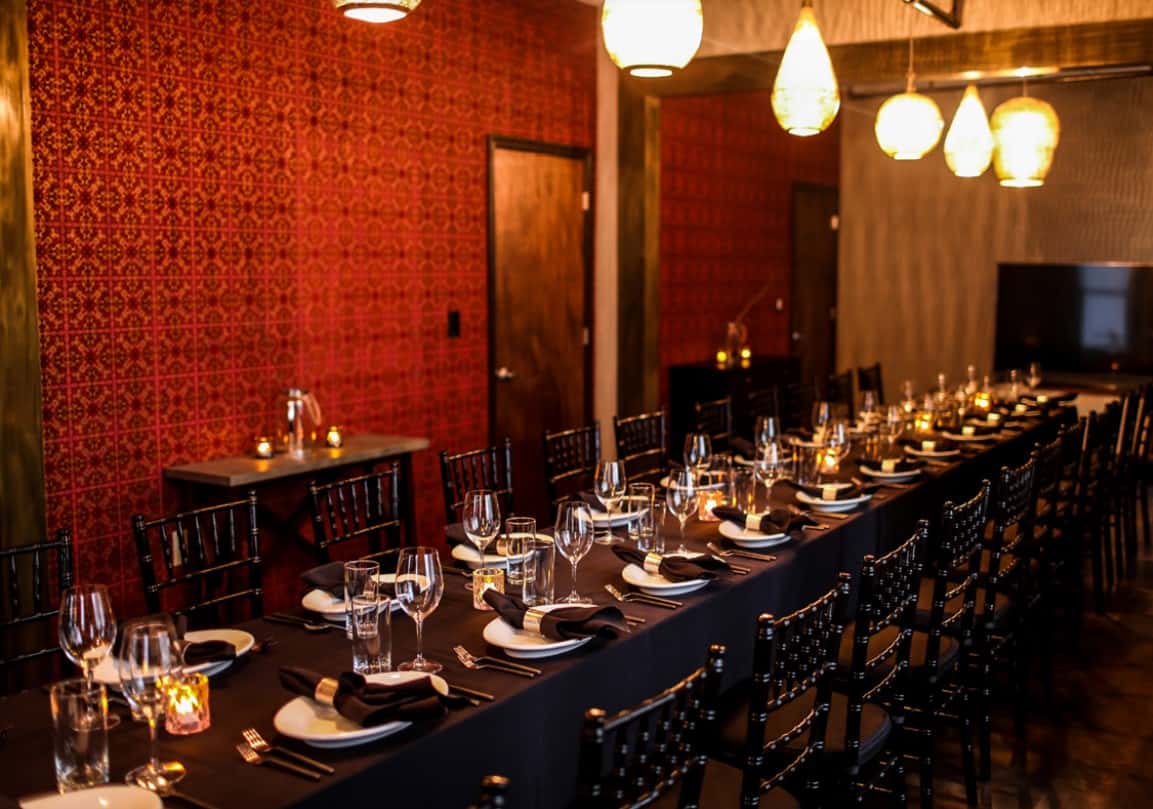 A long dining table with glassware, black napkins, and candles, set against a red patterned wall with soft lighting.