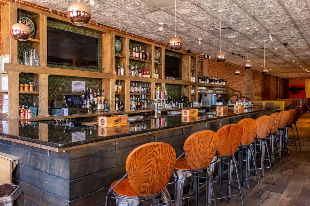 The image shows a modern bar area with a large wooden counter, several high stools, and shelves lined with various bottles of liquor. The bar has stylish overhead lights and a rustic interior with a brick wall.