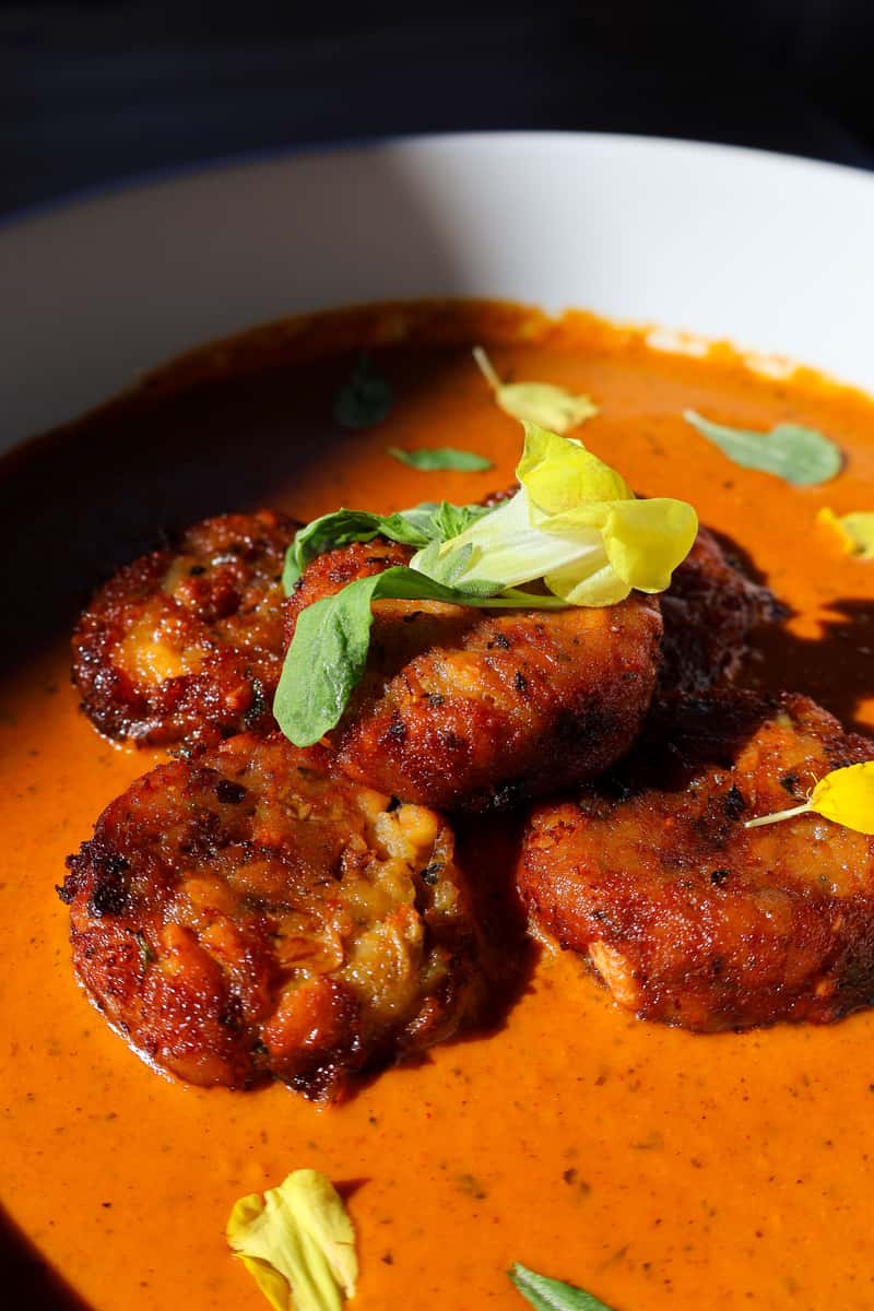 A close-up shot of a creamy orange curry with golden-brown, crispy meatballs, garnished with fresh herbs and colorful edible flowers.