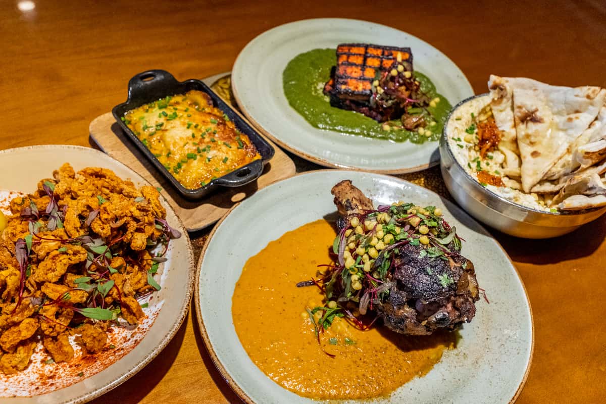 A selection of five different dishes, including a grilled meat with a spiced sauce, a serving of crispy fried food, a cheesy casserole, naan, and a creamy side dish.