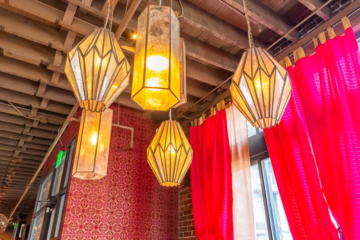 A set of pendant lights with geometric designs hanging from a wooden ceiling, with bright red curtains in the background.
