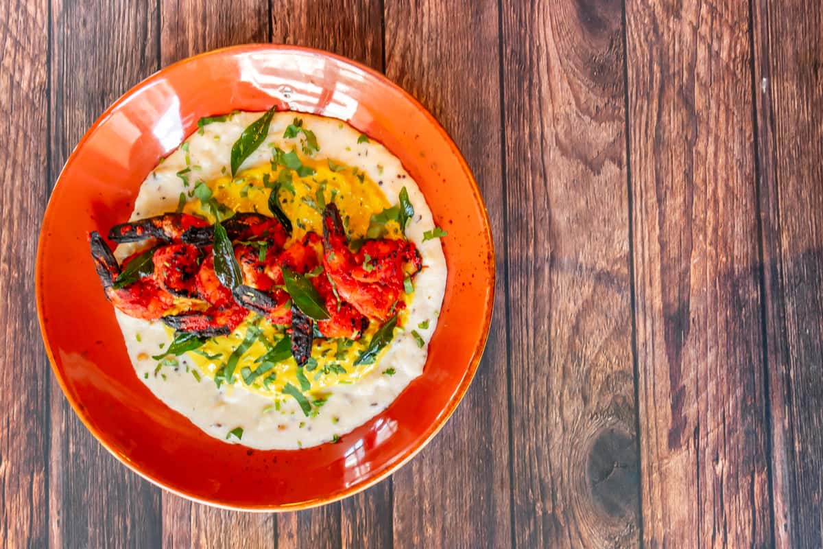 A plate with grilled shrimp placed on a creamy, yellow sauce garnished with fresh herbs and spices. The plate has an orange rim, set on a wooden table.