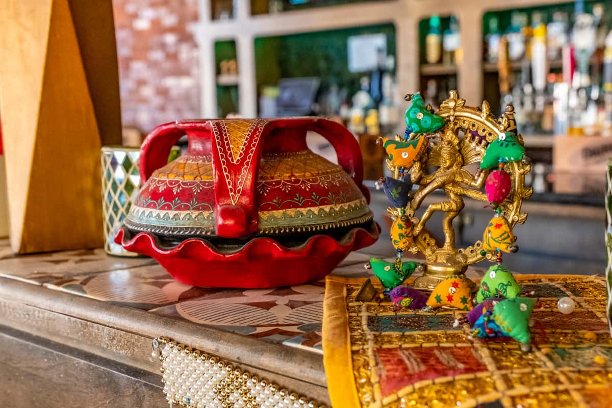 The image shows a colorful traditional display with a red pottery vessel, a golden statue of a dancer, and decorative fabric items, set on a decorated surface.