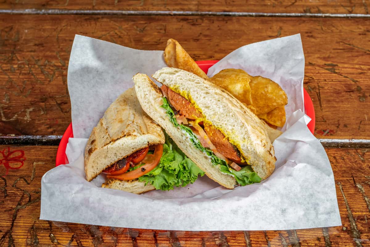 Toasted sandwich on a hoagie roll filled with grilled sausage, fresh lettuce, tomato, onions, and mustard, served with crispy kettle chips in a red plastic basket lined with parchment paper on a rustic wooden table.