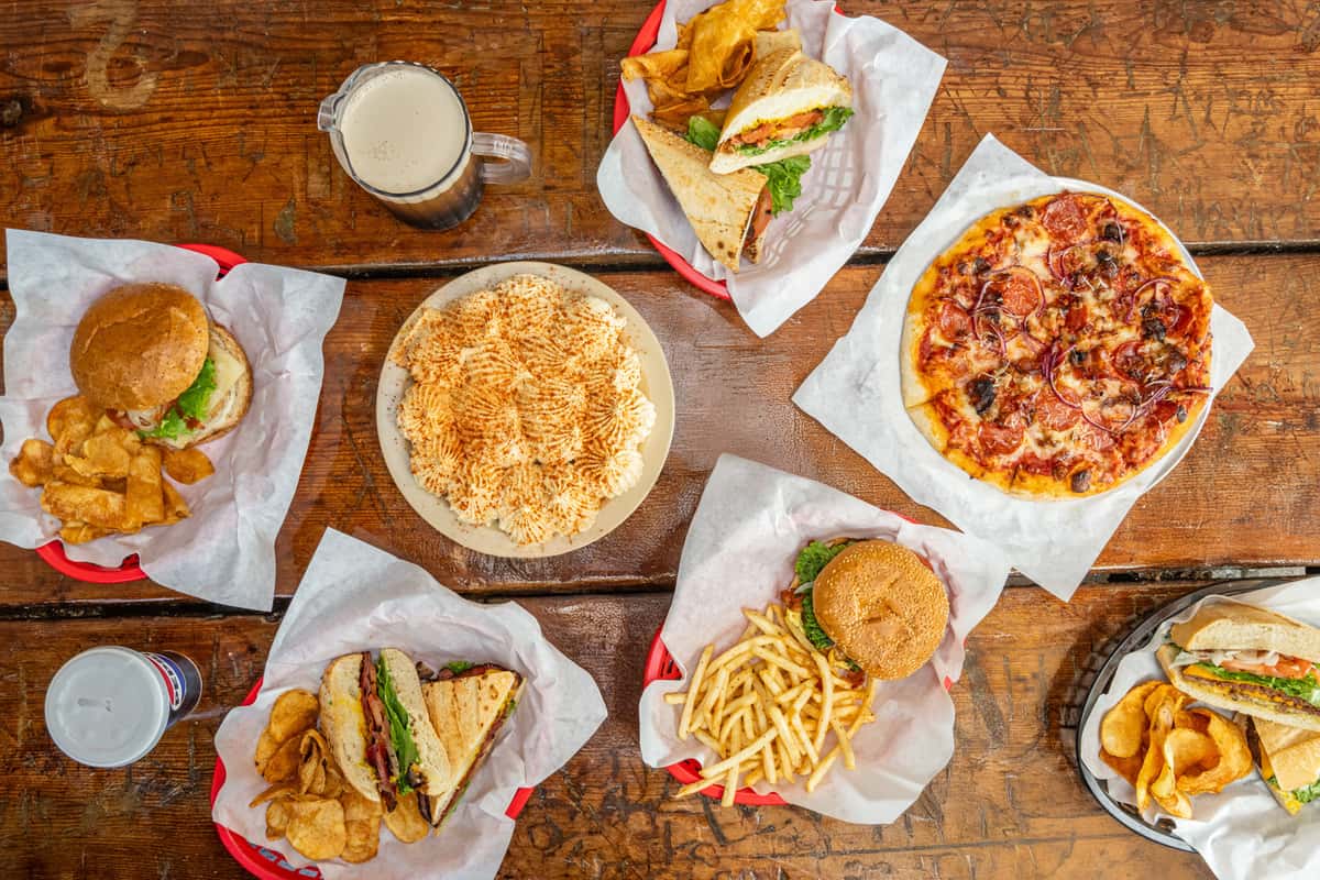 Overhead view of a casual dining spread on a rustic wooden table, featuring a mix of hearty American comfort foods including burgers with fries and kettle chips, deli sandwiches with fresh veggies, a loaded pepperoni and red onion pizza, a cheesy baked casserole topped with paprika, and a glass of dark beer alongside a soft drink.