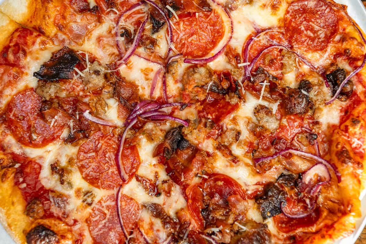 Close-up of a freshly baked meat lover's pizza topped with pepperoni, crumbled sausage, crispy bacon, red onions, and melted mozzarella cheese, all on a golden-brown crust with a zesty tomato sauce base.