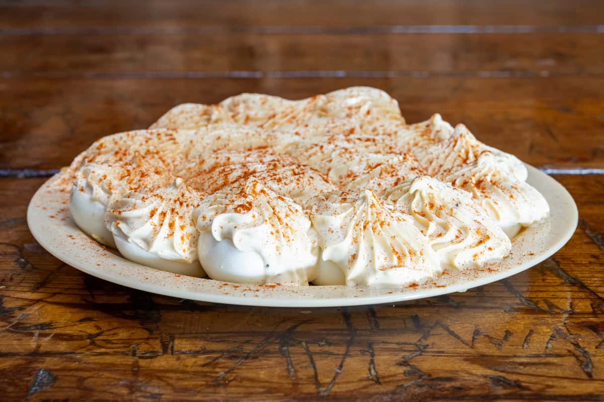 Plate of deviled eggs piped with a creamy, seasoned filling and dusted with paprika, arranged in a circular pattern on a ceramic dish over a rustic wooden table.