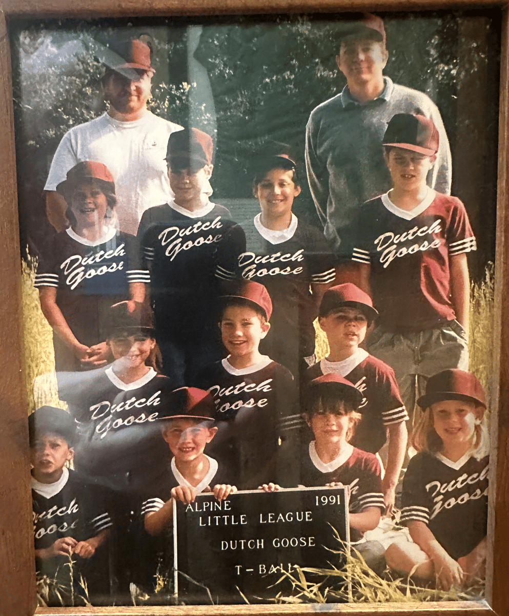 Youth baseball team in matching Dutch Goose uniforms poses outdoors with coaches, holding an Alpine Little League T-Ball sign.