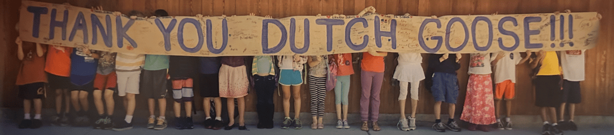 Children stand side by side holding a handmade banner reading "Thank You Dutch Goose!!!" with colorful clothing visible below the sign