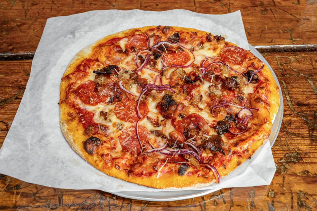 Freshly baked meat lover's pizza topped with pepperoni, sausage, bacon, red onions, and melted mozzarella cheese, served on parchment paper over a metal tray atop a rustic wooden table.