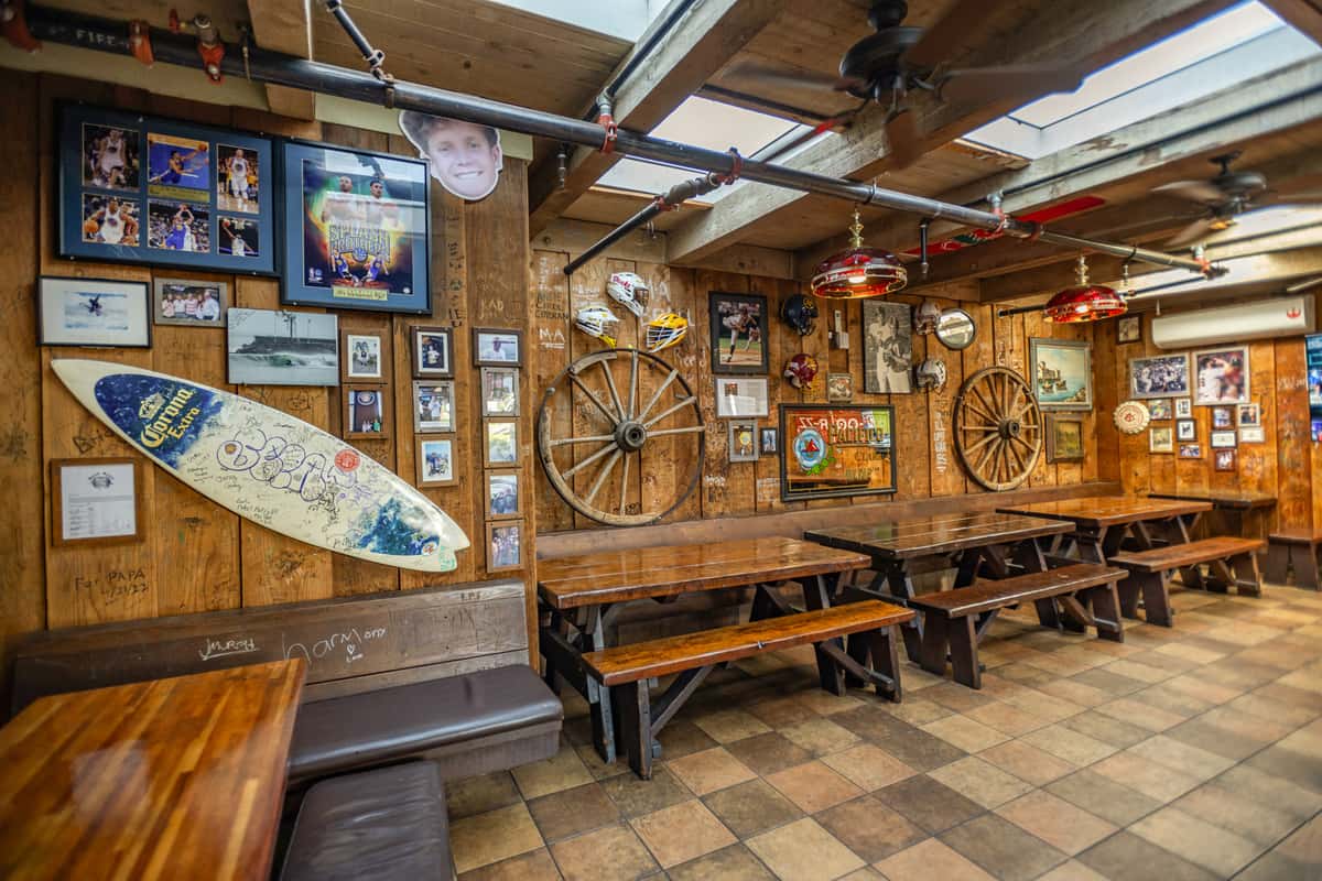 Rustic dining area with wooden picnic-style tables and benches, surrounded by wood-paneled walls adorned with eclectic memorabilia, including framed sports photos, vintage signs, a Corona-branded surfboard, and old wagon wheels. Overhead, exposed piping and retro pendant lights enhance the cozy, nostalgic bar-and-grill vibe.