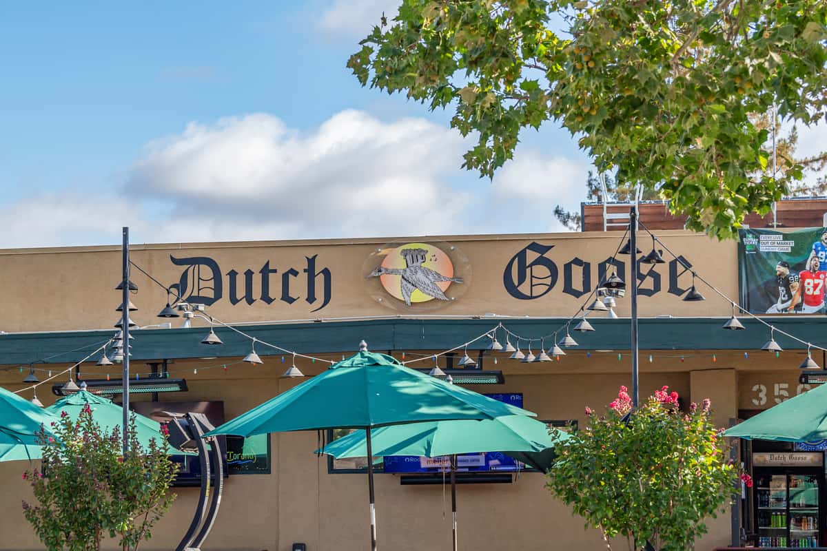 Exterior of Dutch Goose restaurant featuring bold vintage-style signage, green patio umbrellas, string lights, and flat-screen TVs, creating a laid-back, sports bar atmosphere under a clear blue sky.