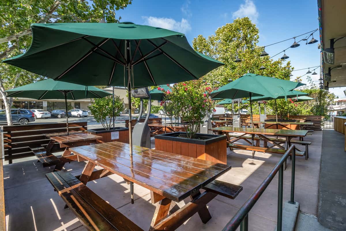 Spacious outdoor dining patio with polished wooden picnic tables, large green umbrellas for shade, and potted flowering plants, set along a tree-lined street with string lights overhead—ideal for casual, al fresco dining.
