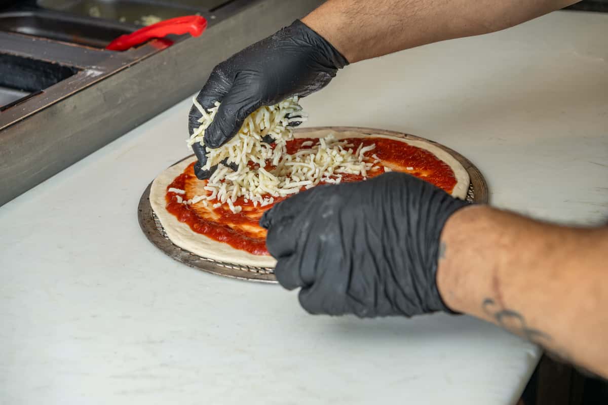 Close-up of a cook wearing black gloves sprinkling shredded mozzarella cheese over a pizza crust topped with tomato sauce, preparing a fresh pie on a metal tray in a commercial kitchen setting.