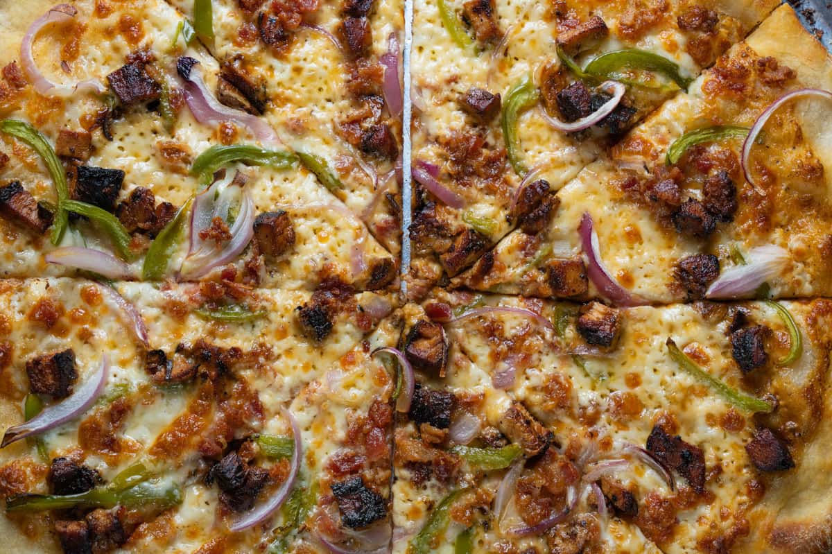 Close-up of a specialty pizza topped with melted mozzarella, crispy bacon or grilled meat chunks, green bell peppers, and sliced red onions on a golden hand-tossed crust – hearty gourmet pizza perfect for a casual Italian restaurant or neighborhood pizzeria menu.
