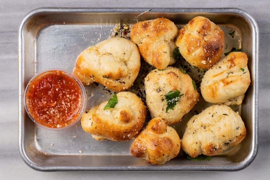 Garlic Parmesan knots baked to golden perfection and sprinkled with herbs, served in a metal tray with a side of marinara dipping sauce – classic Italian appetizer perfect for a casual pizzeria or family-style restaurant menu.