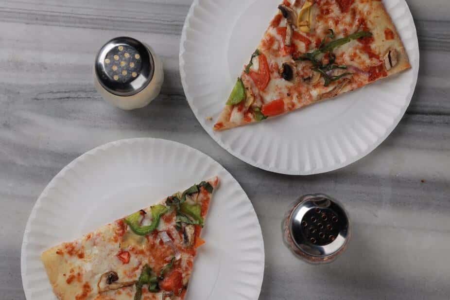 Two slices of vegetarian pizza topped with bell peppers, red onions, mushrooms, and diced tomatoes served on white plates, with grated Parmesan and red pepper flakes shakers on a marble table – casual pizzeria dining scene perfect for an Italian restaurant menu or lunch special promotion.