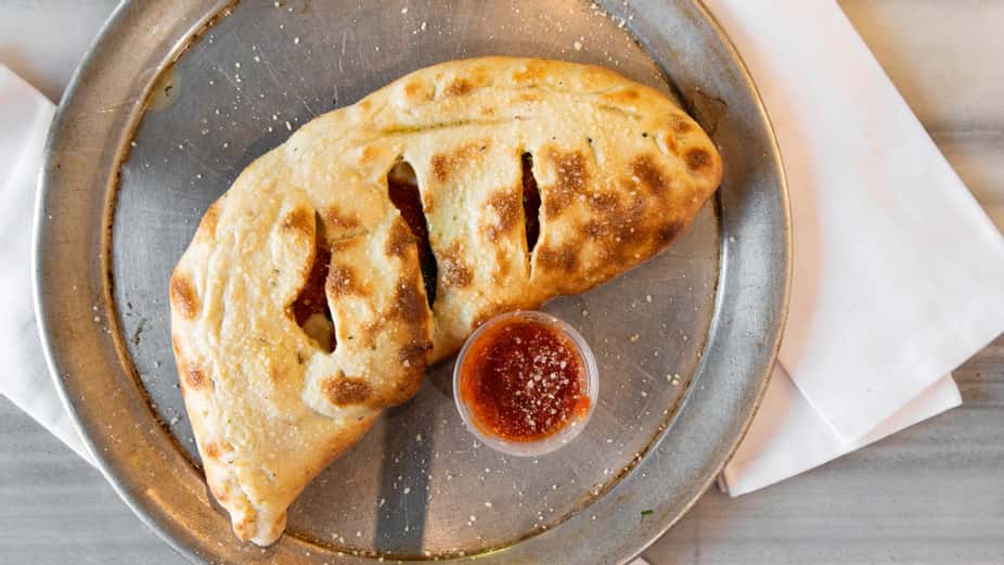 Oven-baked calzone stuffed with melted cheese and savory fillings, served with a side of marinara dipping sauce on a metal tray – golden, folded Italian pizza pocket perfect for a casual pizzeria menu.
