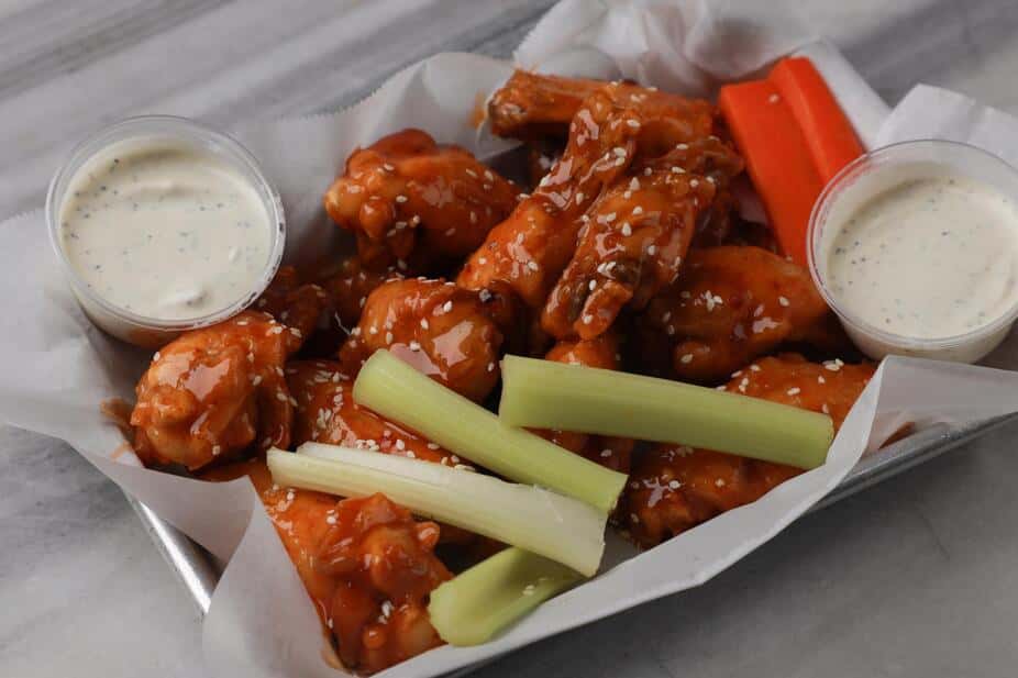 Buffalo chicken wings coated in tangy hot sauce and sprinkled with sesame seeds, served in a parchment-lined tray with crisp celery and carrot sticks and creamy ranch dipping sauce – classic spicy bar food perfect for a sports bar or casual American restaurant menu.