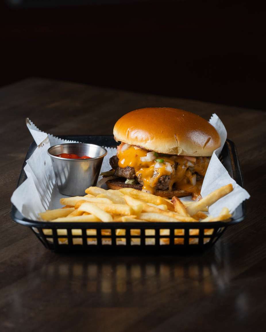 Juicy cheeseburger topped with melted cheddar and grilled onions on a toasted brioche bun, served with crispy French fries and a side of ketchup in a basket – classic American diner burger perfect for a casual restaurant or sports bar menu.