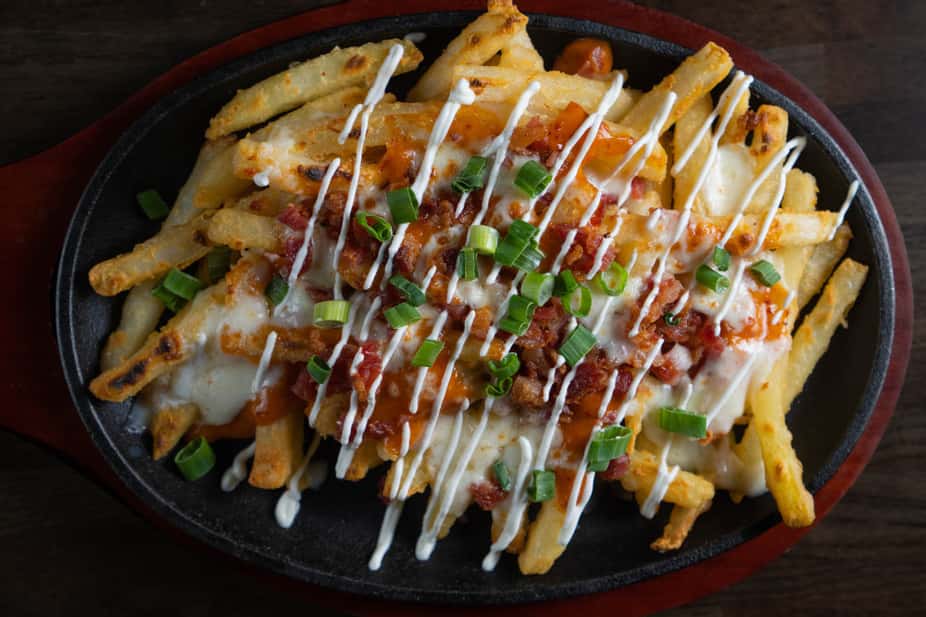 Loaded fries topped with melted cheese, crispy bacon bits, drizzled ranch and spicy buffalo sauce, and fresh chopped green onions, served in a cast iron skillet – indulgent comfort food appetizer perfect for a casual American restaurant or sports bar atmosphere.