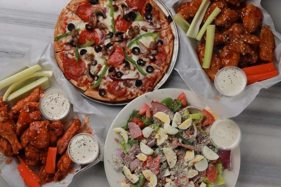 Overhead view of a supreme pizza topped with pepperoni, sausage, olives, peppers, and onions, served alongside buffalo chicken wings with celery sticks and ranch dressing, plus a fresh chef salad with hard-boiled eggs – complete Italian-American pizzeria feast perfect for family dinner or game day spread.