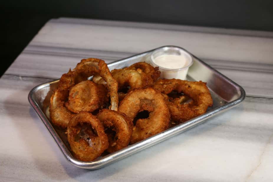 Crispy golden onion rings served in a metal tray with a side of creamy ranch dipping sauce – classic bar appetizer perfect for a casual American restaurant or sports bar menu.