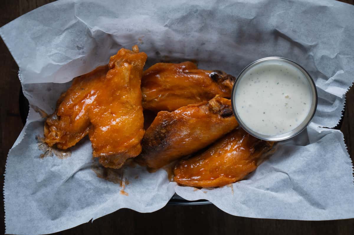 Buffalo chicken wings coated in tangy orange hot sauce, served in a parchment-lined basket with a side of creamy ranch dipping sauce – classic spicy bar appetizer perfect for a sports bar or casual American restaurant menu.