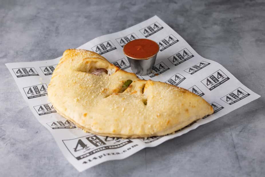 Golden-brown baked calzone filled with melted cheese and savory pizza toppings, served on branded parchment paper with a side of marinara dipping sauce – classic Italian pizzeria favorite perfect for a casual restaurant menu.
