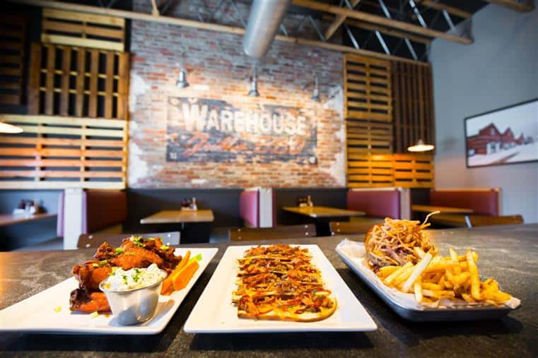 Warehouse Grill and Barbecue dining room with booths. Wings, pulled pork nachos, and pulled pork sandwich with fries on three dishes in foreground