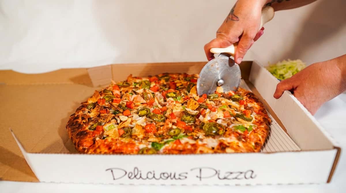 Pizza with jalapeños, diced tomatoes, and ground meat being sliced with a pizza cutter in a box.